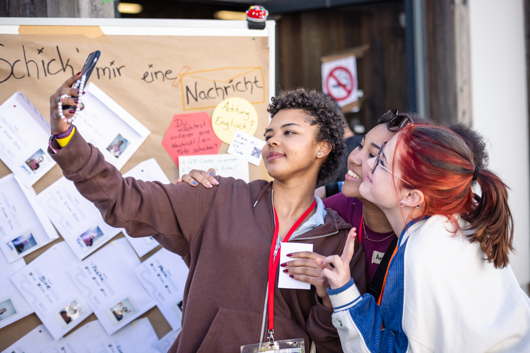 Drei weiblich gelesene Personen stehen beisammen vor einer dekorierten Pinnwand und machen zusammen ein Selfie. ©Servicestelle Jugendarbeit 2022