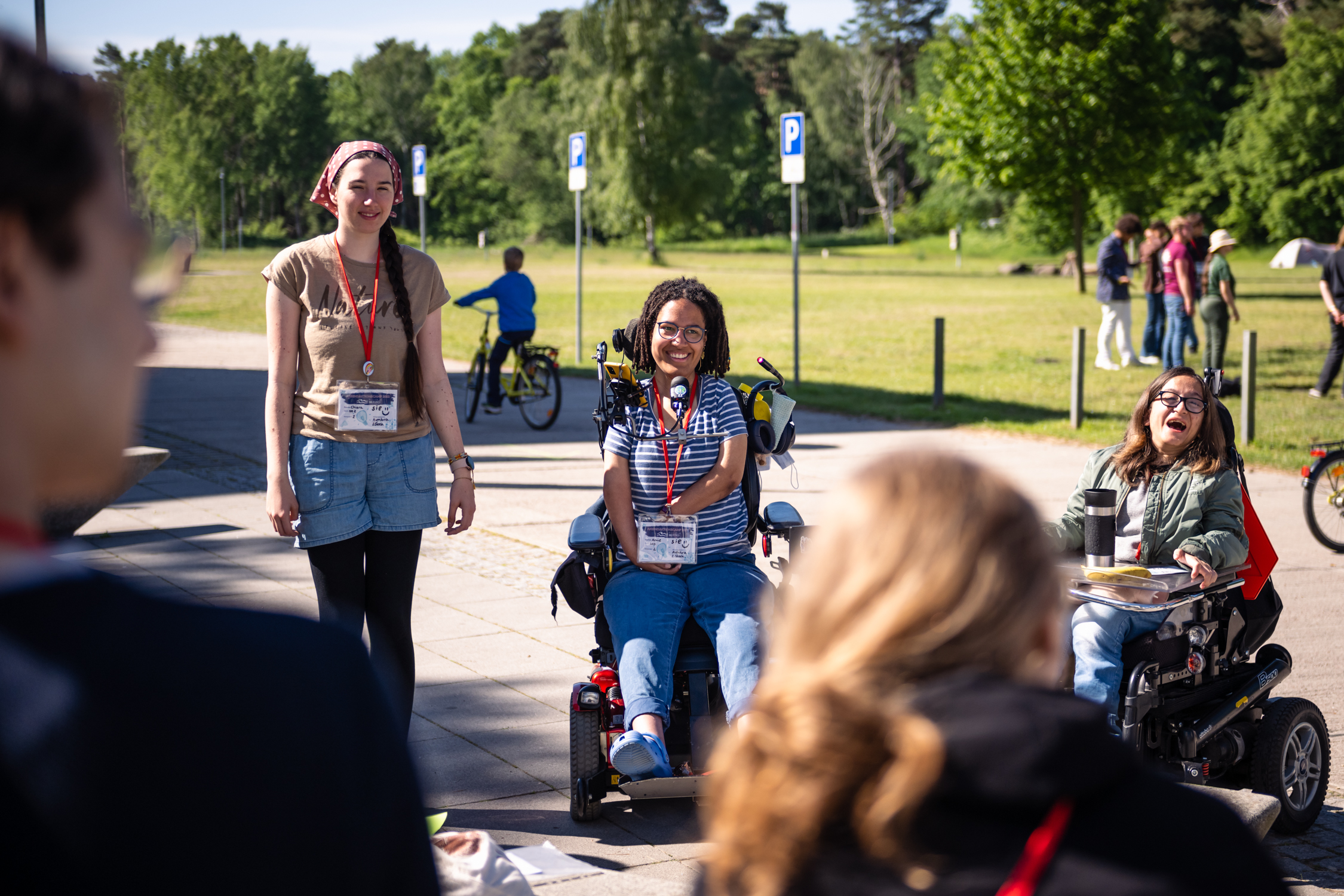 Es sind drei junge Frauen vom JAC 2022 abgebildet. Links steht eine Frau und rechts sitzen die zwei weiteren Frauen im Rollstuhl. Sie alle lächeln in die Kamera. ©Servicestelle Jugendbeteiligung 2022