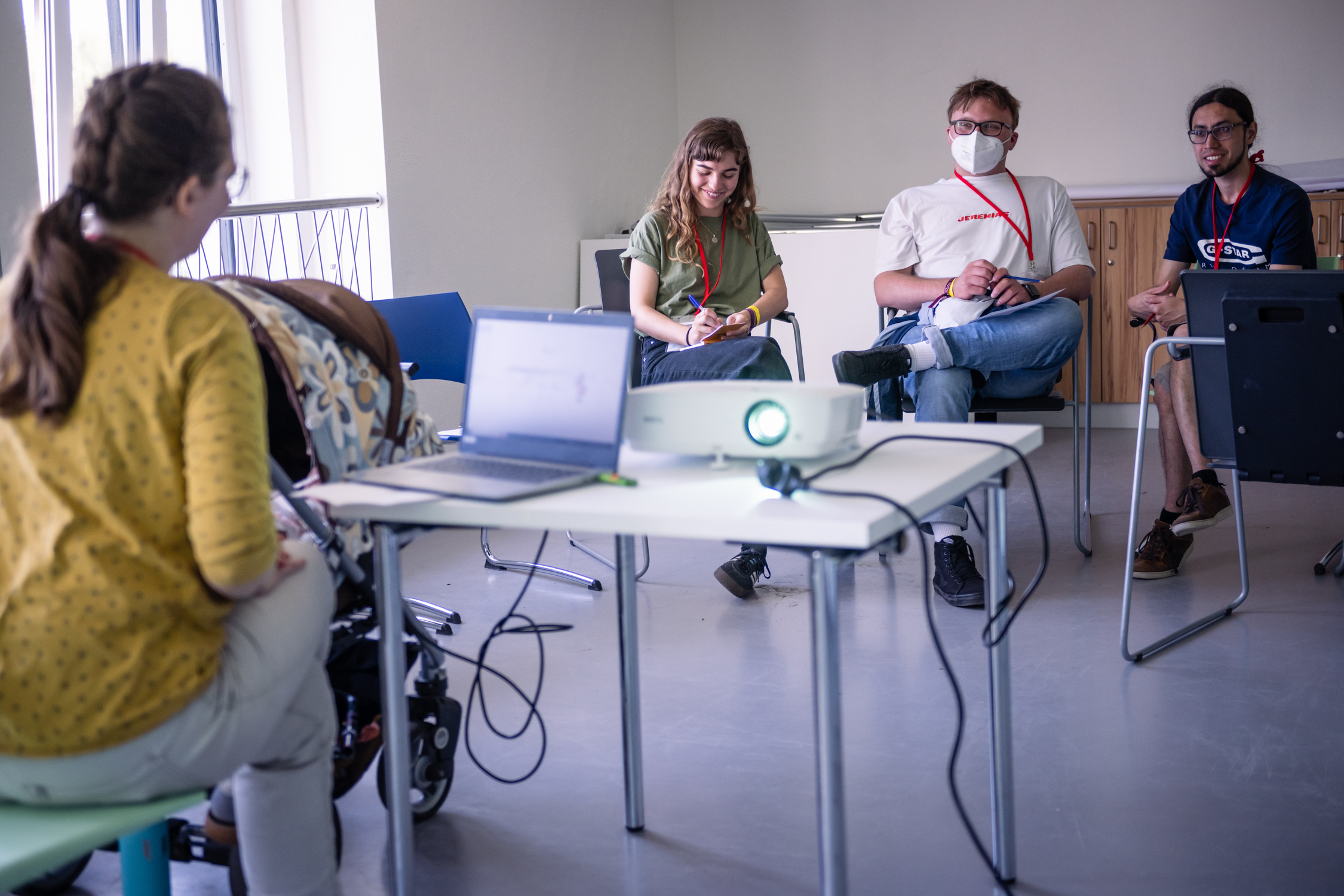 Es handelt sich um eine Aufnahme von einem Workshop während des JACs 2022. Eine Frau leitet den Workshop, ist mit dem Rücken zur Kamera und arbeitet mit ihrem Laptop an einem Tisch an dem sie sitzt. Ihr gegenüber sitzen drei Teilnehmende, die ihr zuhören. ©Servicestelle Jugendbeteiligung 2022