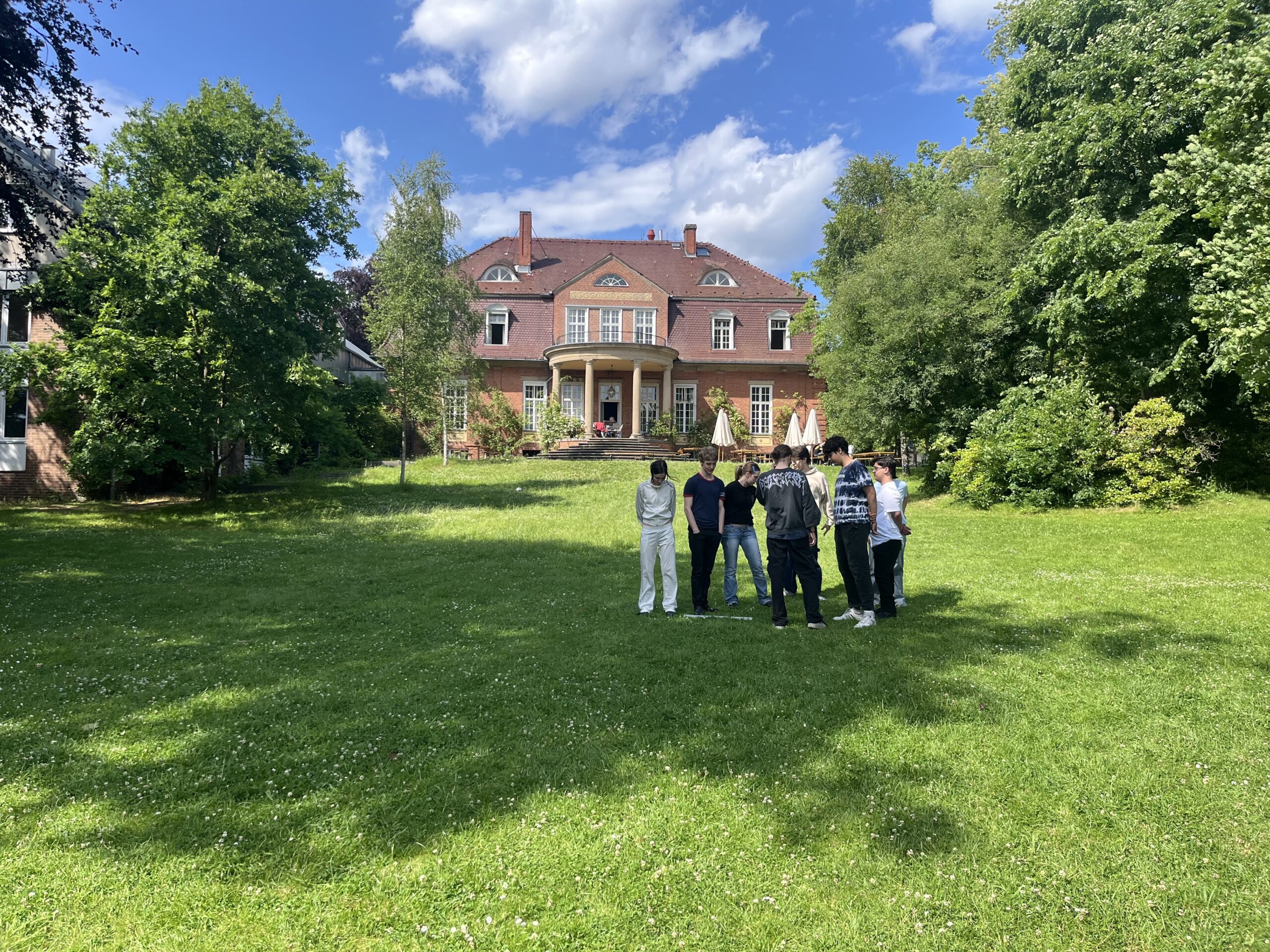 Auf eine großen sonnenbeschienenen Wiese steht eine Gruppe von 7 jungen Erwachsenen im kreis und bespricht etwas. Im Hintergrund steht ein großes schickes Haus, das wannseeFORUM